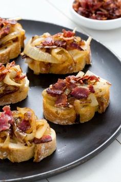 four pieces of bread topped with bacon and onions on a black plate next to a white bowl