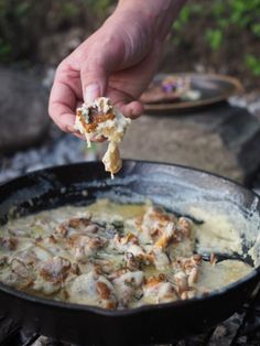 a person is dipping some food into a skillet on the campfire with other items nearby