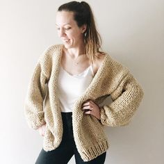 a woman standing in front of a white wall wearing a cardigan sweater and black pants