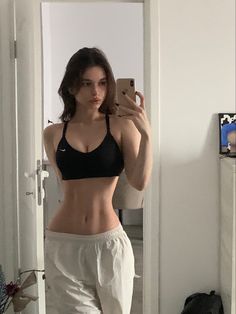 a woman taking a selfie in the mirror wearing white pants and a black bra top