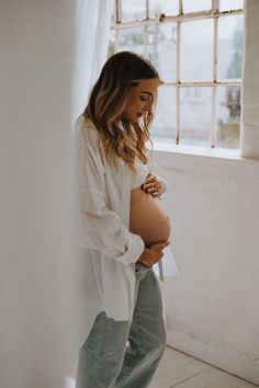 a pregnant woman standing in front of a window with her belly tucked under her shirt