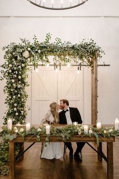 a bride and groom kissing in front of a wooden table with greenery on it