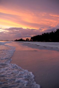 the sun is setting at the beach with waves coming in to shore and trees lining the shoreline