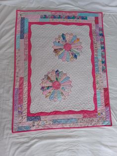 a pink and blue quilt on a white bed with two flowers in the center,