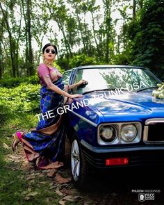 a woman leaning on the hood of a blue car in front of trees and grass