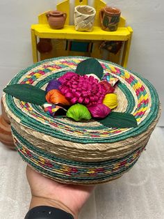 a hand holding a basket with yarn and flowers in it on top of a table