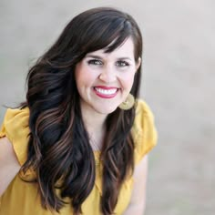 a woman with long dark hair wearing a yellow top and gold earrings smiling at the camera