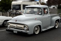 an old gray truck driving down the road next to a white car and a trailer