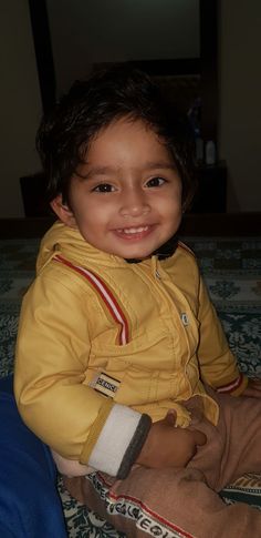 a little boy sitting on top of a bed wearing a yellow jacket and smiling at the camera