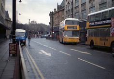 two double decker buses are parked on the side of the street as people walk by