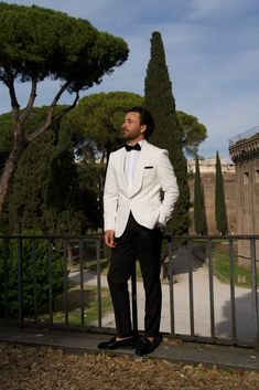 a man in a white tuxedo standing next to a fence and looking off into the distance