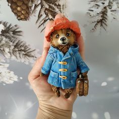 a hand holding a teddy bear wearing a blue coat and orange hat with pine cones in the background