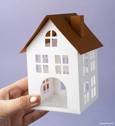 a hand holding up a paper model of a white house with brown roof and windows