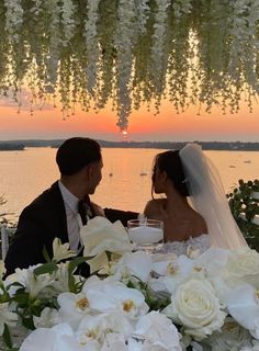 a bride and groom sitting at a table with flowers in front of the sun setting