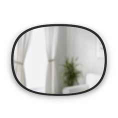 the reflection of a white chair in a black oval mirror with curtains and a potted plant behind it