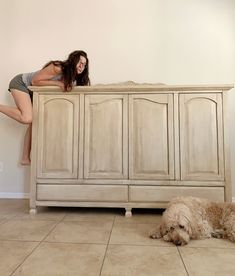 a woman standing on top of a dresser next to a dog laying on the floor