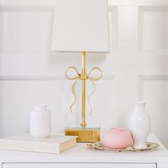 a table with a lamp, vases and a book on top of it in front of a white wall