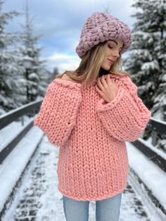 a woman standing in the snow wearing a pink knitted sweater and beanie hat