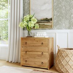 a wooden dresser sitting next to a window with white flowers on it and a painting hanging above