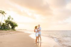 a man and woman standing on top of a beach next to the ocean at sunset