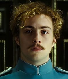 a man with curly hair and a moustache looks at the camera in front of bookshelves