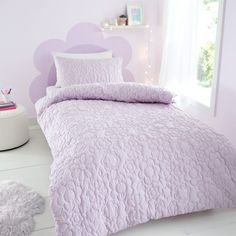 a bed with purple comforter and pillows in a pink room next to a window