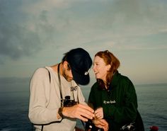 a man and woman standing next to each other on top of a cliff near the ocean