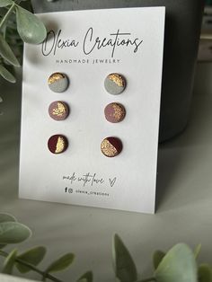 three pairs of earrings with gold and red accents on a card next to some plants