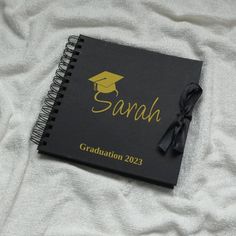 a black notebook with yellow lettering and a graduation cap on the cover is laying on a white blanket