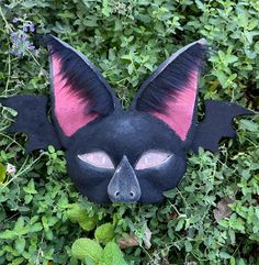 a black cat mask with pink eyes and ears laying in the grass next to green plants