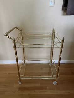 a glass and brass serving cart with wheels on the floor in front of a wall