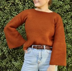 a woman standing in front of a hedge wearing a brown sweater and jeans with her hands on her hips