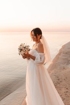 a woman in a wedding dress standing on the beach holding a bouquet of white flowers