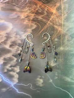 This enchanting mermaid moon ear cuff is sure to make a statement and bring out your inner magic! Silver ear cuff is embellished with a charm of two women in the moon, rainbow mermaid tail charm, iridescent drop crystals, seashell and starfish, and electroplated lava stone beads all dangling from silver and rainbow chains. This ear cuff is perfect for adorning pierced and non-pierced ears, and is lightweight and comfortable to wear. Lava Stone offers the following positive energies for the weare Unique Iridescent Jewelry For Festivals, Whimsical Adjustable Ear Cuff For Festivals, Bohemian Iridescent Jewelry With Ear Wire, Bohemian Dangle Body Jewelry For Festivals, Nickel-free Fantasy Jewelry For Festivals, Bohemian Metal Dangle Body Jewelry, Fantasy Style Festival Ear Cuff, Fantasy Dangle Jewelry For Festivals, Fantasy Style Festival Ear Cuff For Pierced Ears