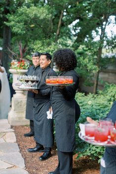 two waiters holding trays of food in front of some other people and trees