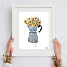 two hands holding up a framed print with flowers in a watering can on the table