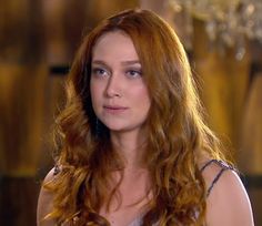 a woman with long red hair sitting in front of a chandelier and looking at the camera