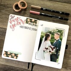 an open wedding album on a wooden table next to some pens and pencils, with a couple holding each other's hands