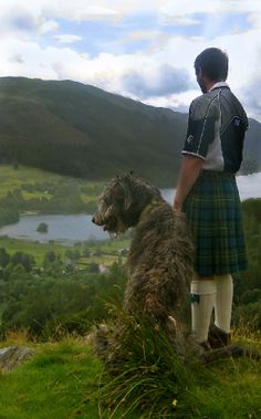 a man standing on top of a lush green hillside next to a dog wearing a kilt