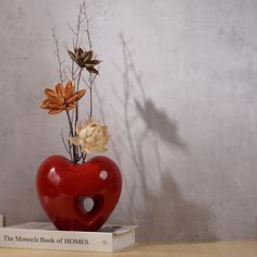 a red heart shaped vase sitting on top of a book next to a flower arrangement