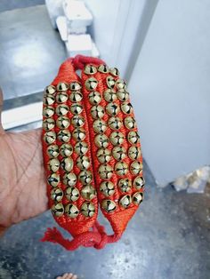 a hand holding two red bracelets with gold bells on it's ends and one has an orange ribbon around the end