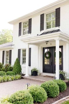 a white house with black shutters and wreath on the front door is surrounded by greenery