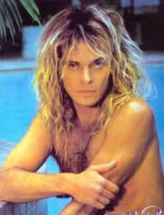 a man with long blonde hair sitting next to a swimming pool and posing for the camera