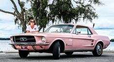 a woman sitting on the hood of a pink mustang parked in front of a body of water