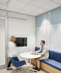 a man sitting at a white table in an office with blue chairs and a television