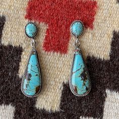 These genuine turquoise and sterling silver handmade dangle earrings are a harmonious blend of elegance and artisan craftsmanship. The focal point of each earring is a mesmerizing turquoise gemstone, displaying its natural beauty with varying shades of greenish blue and hints of earthy matrix, making each piece unique. The turquoise stones are carefully set within sterling silver bezels, enhancing their allure and providing a stunning contrast against the silver backdrop. The intricate detailing of the earrings includes rope borders that gracefully encircle the turquoise stones, adding a touch of texture and sophistication to the design. The dangle style allows the earrings to sway delicately with movement, catching the light and drawing attention to the exquisite craftsmanship. At the top Southwestern Turquoise Drop Earrings, Southwestern Turquoise Earrings With Natural Stones, Turquoise Southwestern Earrings With Natural Stones, Silver Backdrop, Handmade Dangle Earrings, Faith Jewelry, Earrings Handmade Dangle, Earrings Turquoise, Greenish Blue