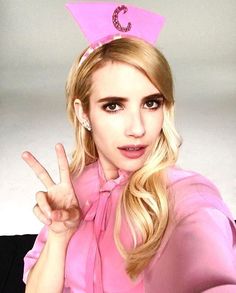 a woman with blonde hair wearing a pink top and holding up her peace sign