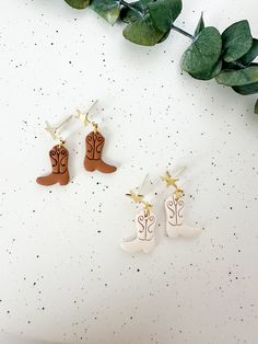 three pairs of cowboy boots are hanging from earrings on a white surface next to green leaves