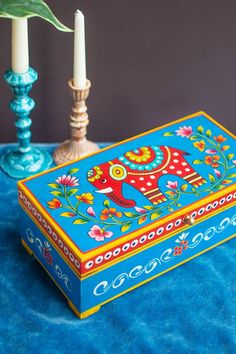 a blue box with an elephant painted on it next to two candles and a green plant