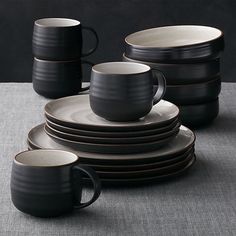 a stack of black and white dishes on a gray tablecloth with two mugs in the middle